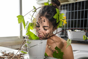 Frau bedeckt mit Pflanzenblatt riechenden Topfpflanze Schlamm zu Hause - GGGF00215