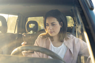 Junge Frau, die im Auto sitzend ihr Smartphone benutzt, während sie bei Sonnenuntergang unterwegs ist - AFVF07590