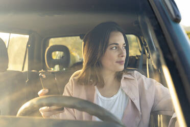 Frau mit Handy in der Hand, die während einer Autofahrt bei Sonnenuntergang wegschaut - AFVF07591