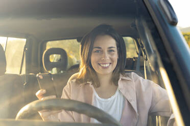 Lächelnde junge Frau mit Handy im Auto bei Sonnenuntergang - AFVF07592