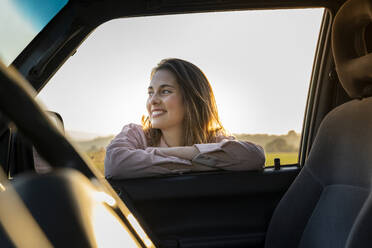 Glückliche junge Frau, die sich bei Sonnenuntergang an ein Autofenster lehnt und wegschaut - AFVF07595