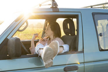 Junge Frau, die wegschaut, während sie mit hochgelegten Füßen auf dem Autofenster während einer Autoreise sitzt - AFVF07601