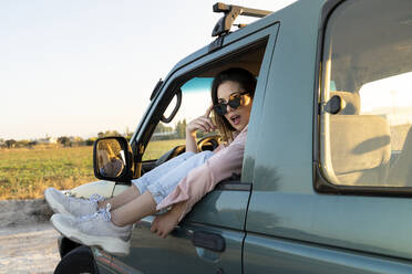 Erstaunte junge Frau, die während einer Autofahrt bei Sonnenuntergang mit den Füßen auf dem Autofenster wegschaut - AFVF07604