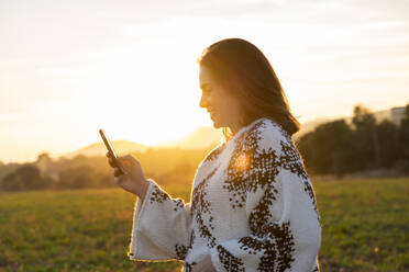 Lächelnde junge Frau, die bei Sonnenuntergang ein Mobiltelefon benutzt - AFVF07612