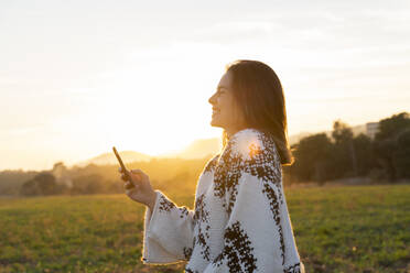 Fröhliche junge Frau, die während des Sonnenuntergangs ein Mobiltelefon benutzt - AFVF07613