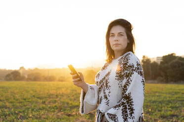 Schöne junge Frau, die ihr Handy hält und bei Sonnenuntergang wegschaut - AFVF07614