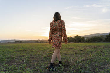 Junge Frau in geblümtem Sommerkleid geht bei Sonnenuntergang auf einer Wiese spazieren - AFVF07618