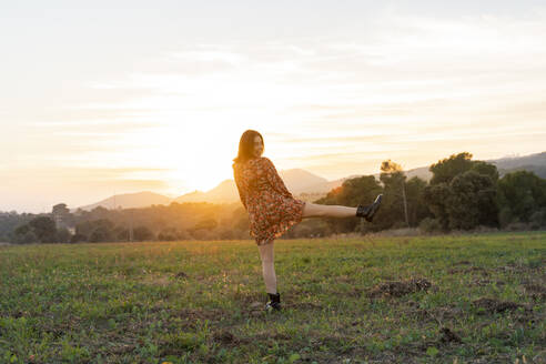 Fröhliche junge Frau auf einem Bein stehend auf einer Wiese bei Sonnenuntergang - AFVF07620