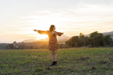 Young woman in sundress with arms outstretched against sky during sunset - AFVF07621