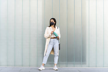 Female entrepreneur with bottle standing on footpath against glass wall during COVID-19 - AFVF07635