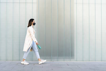 Businesswoman with bottle walking on footpath by glass wall during pandemic - AFVF07636
