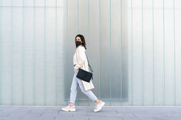 Female entrepreneur walking on footpath by glass wall during coronavirus - AFVF07637