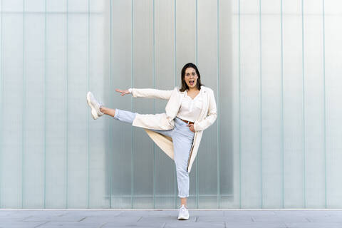 Cheerful businesswoman with stretching leg and arm while standing against glass wall stock photo