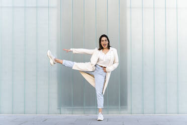 Cheerful businesswoman with stretching leg and arm while standing against glass wall - AFVF07639