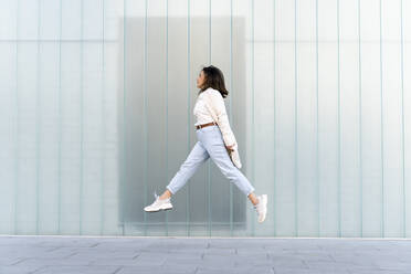 Businesswoman jumping on footpath by glass wall - AFVF07643