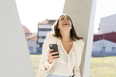 Female entrepreneur laughing while holding smart phone in city on sunny day - AFVF07656