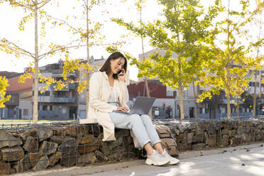 Female entrepreneur talking through smart phone while using laptop sitting on retaining wall - AFVF07661