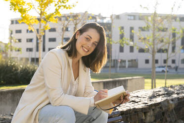 Lächelnde Geschäftsfrau mit Tagebuch auf einer Stützmauer sitzend - AFVF07666