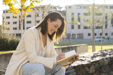 Lächelnde Geschäftsfrau, die auf einer Mauer sitzend in ihr Tagebuch schreibt - AFVF07667