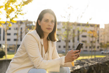 Businesswoman holding smart phone looking away while sitting in city - AFVF07671