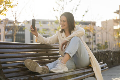 Lächelnde Geschäftsfrau, die ein Selfie mit ihrem Smartphone macht, während sie auf einer Bank sitzt, lizenzfreies Stockfoto