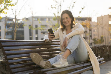 Happy businesswoman holding smart phone while sitting on bench - AFVF07674