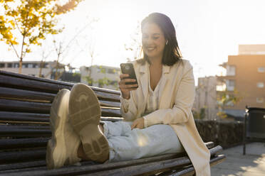 Unternehmerin lächelt, während sie auf einer Bank sitzend ein Smartphone benutzt - AFVF07675