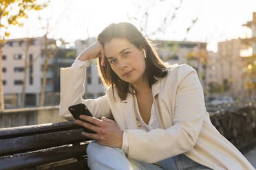 Thoughtful female entrepreneur holding smart phone while sitting on bench - AFVF07677