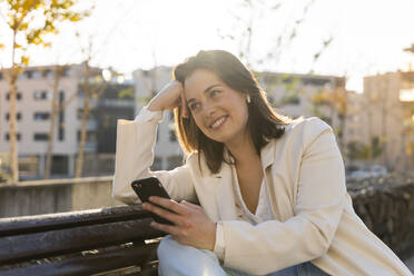 Nachdenklich lächelnde Geschäftsfrau mit Smartphone in der Hand auf einer Bank sitzend - AFVF07678