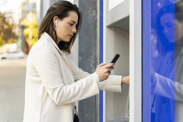 Geschäftsfrau, die ihr Smartphone benutzt, während sie Geld vom Geldautomaten in der Stadt abhebt - AFVF07699