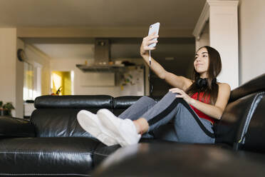 Lächelndes Teenager-Mädchen, das ein Selfie mit seinem Mobiltelefon macht, während es zu Hause auf dem Sofa sitzt - EGAF01080