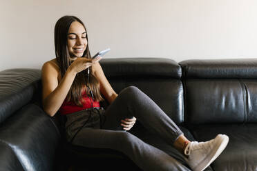 Lächelndes Teenager-Mädchen, das auf dem Sofa zu Hause sitzt und mit dem Handy telefoniert - EGAF01082