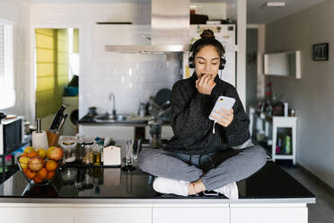 Teenage girl wearing headphones eating fruit while using mobile phone sitting at home - EGAF01092