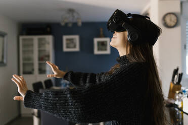 Playful teenage girl wearing virtual reality simulator eyeglasses sitting at home - EGAF01095
