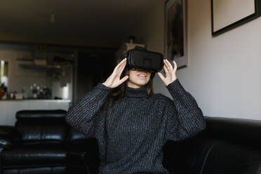 Lächelndes Teenager-Mädchen, das eine Virtual-Reality-Brille benutzt, während es zu Hause auf dem Sofa sitzt - EGAF01097