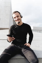 Junger Mann mit Kopfhörern, der lächelnd ein Mobiltelefon benutzt, während er im Freien sitzt - FMOF01214