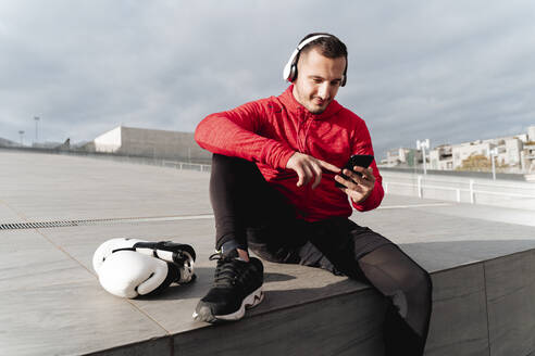 Sportler mit Kopfhörern, der ein Mobiltelefon benutzt, während er im Freien neben Boxhandschuhen sitzt - FMOF01215