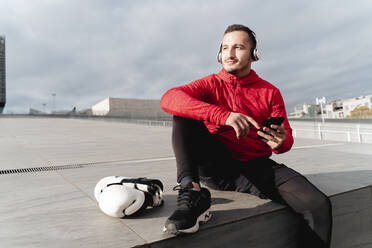 Athlete wearing headphones looking away while using mobile phone sitting outdoors - FMOF01216