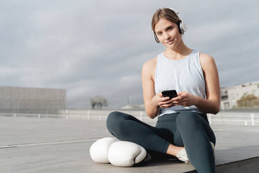 Sportlerin mit Kopfhörern, die im Freien sitzend ein Mobiltelefon benutzt - FMOF01220