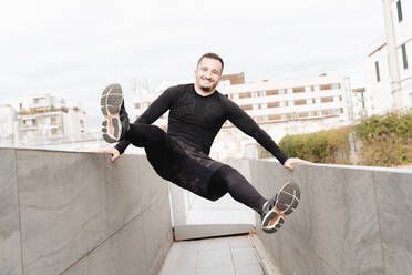 Verspielter Sportler lächelt, während er sich auf der Stützmauer eines Daches schwingt - FMOF01223
