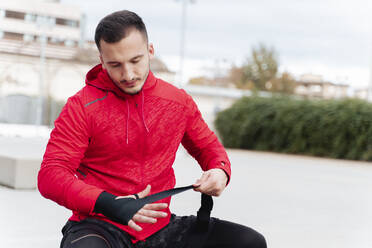 Athlete tying boxing wrap while sitting outdoors - FMOF01232