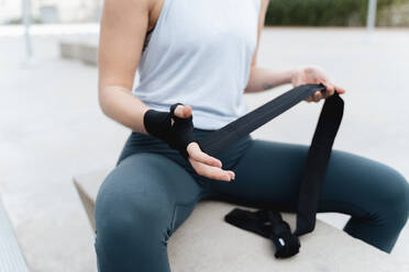 Sportswoman tying boxing wrap in hand while sitting on retaining wall - FMOF01244