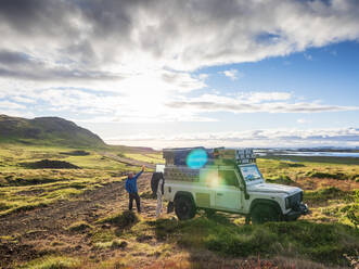 Sun setting over lone traveler standing behind off-road vehicle and waving at camera, Iceland - LAF02594