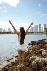 Young female tourist with hands raised enjoying while standing on rocks at bay - MAUF03590