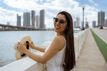 Happy young woman standing at retaining wall while holding hat in city - MAUF03601