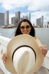 Smiling woman showing hat while standing in city - MAUF03604