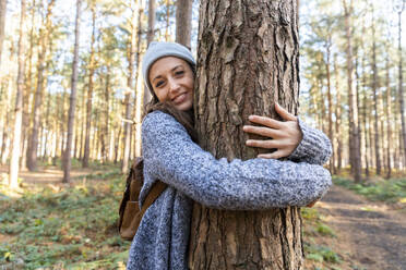 Smiling female hiker embracing tree trunk while exploring in Cannock Chase woodland - WPEF03655
