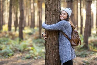 Glückliche Wanderin, die einen Baum umarmt, während sie im Winter im Waldgebiet Cannock Chase wandert - WPEF03658