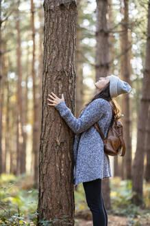 Frau, die im Winter im Wald von Cannock Chase steht und einen Baumstamm betrachtet - WPEF03659