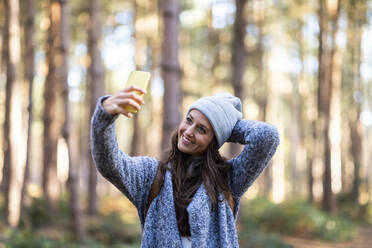Lächelnde Frau mit Hand hinter dem Kopf, die ein Selfie macht, während sie im Wald von Cannock Chase steht - WPEF03668